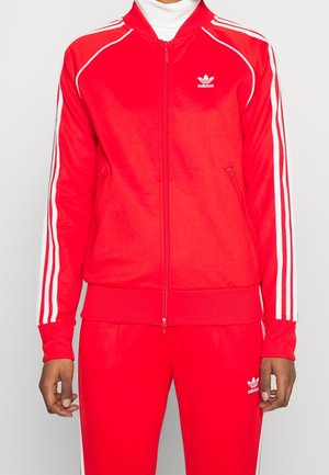 Personne portant un survêtement rouge Adidas avec des bandes blanches sur les manches et les pantalons, et des logos Adidas sur la poitrine et la jambe du pantalon.