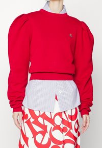 Sudadera roja con mangas abullonadas y dobladillo acanalado, combinada con una camisa a rayas azul y blanca y una falda blanca con letras rojas.