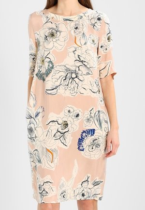 Kobieta ubrana w luźną, sięgającą do kolan brzoskwiniową sukienkę z dużymi abstrakcyjnymi kwiatowymi wzorami w kolorach białym, czarnym, niebieskim i pomarańczowym.