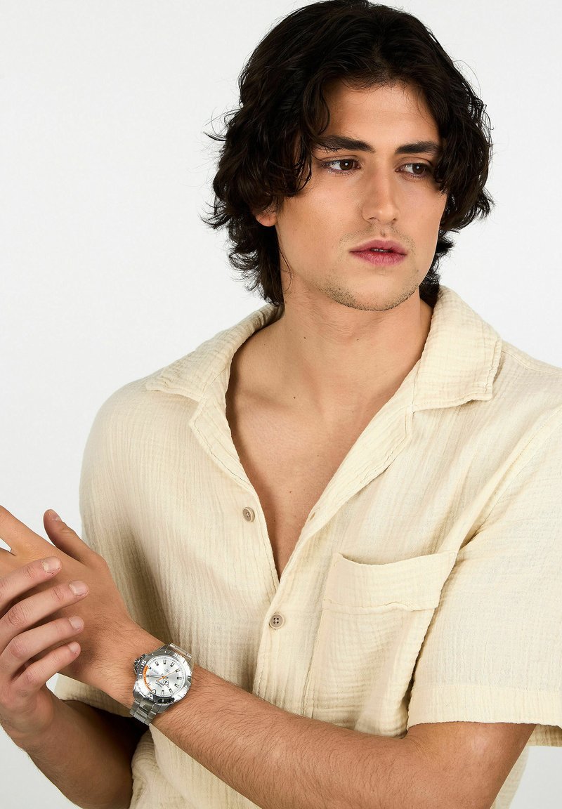 Orologio analogico argento con bracciale in acciaio inossidabile, caratterizzato da un quadrante rotondo, grandi indicatori dell'ora e un accento arancione. Indossato su un polso con una camicia beige.