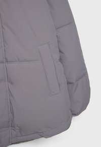 Graue Steppjacke mit matter Oberfläche, ausgestattet mit einer Seitentasche und gesteppten Bereichen zur Isolierung. Weicher, leichter Stoff.