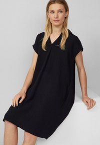 Schwarzes, kurzärmliges Kleid mit lockerer Passform, Kragen, strukturiertem Stoff und seitlichen Taschen, das bis zur Mitte des Oberschenkels reicht.