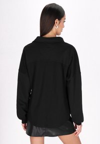 Fekete, túlnőtt ing, textúrált anyagból, gallérral, ejtegetett vállakkal és hátvarrás részletekkel. Simas hemline szoknya felett.