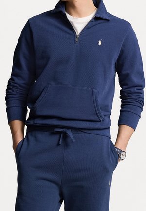 Homme portant un sweat-shirt bleu marine à fermeture éclair quart de zip et un pantalon de survêtement assorti avec logo polo blanc, sous-vêtement blanc visible au niveau du col.