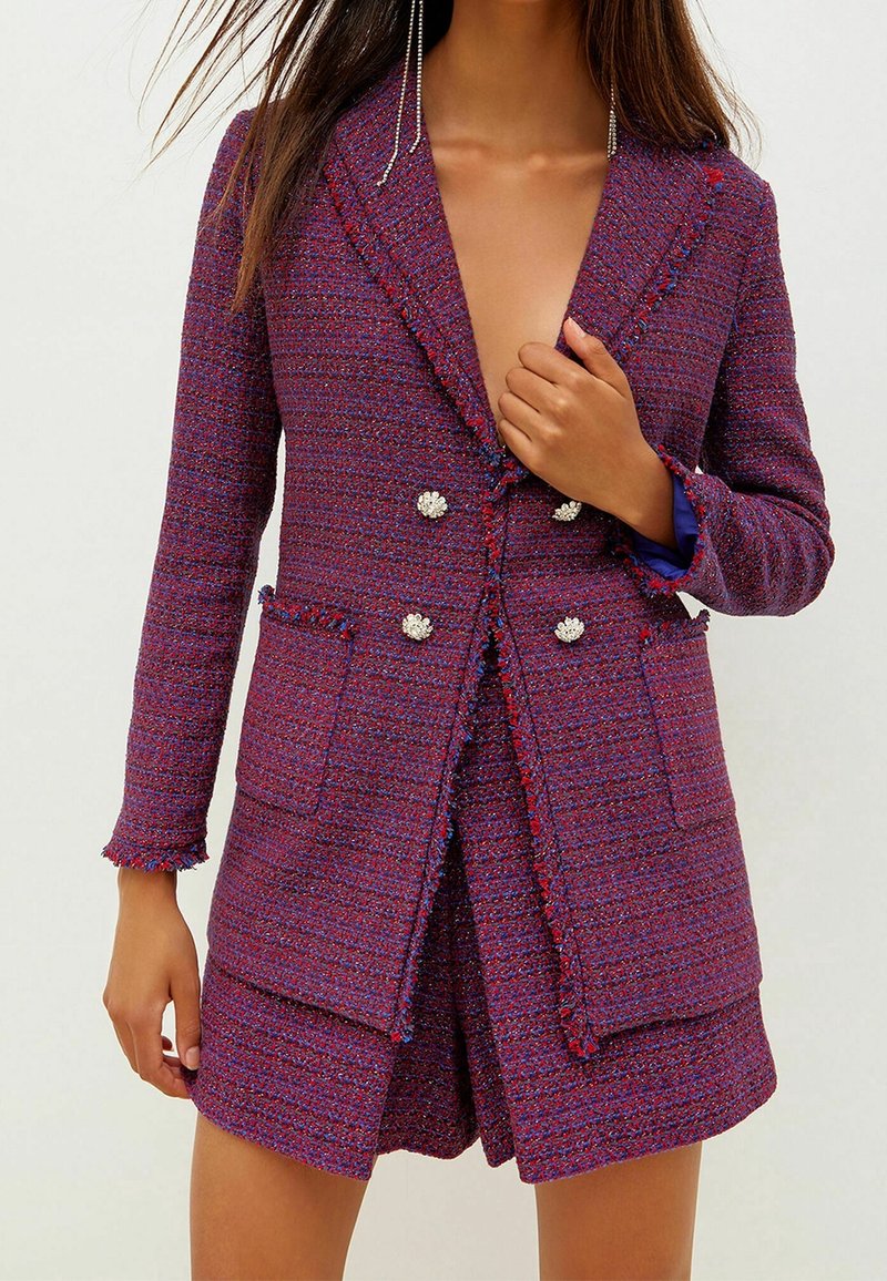 Blazer en tweed violet avec un design croisé, deux poches avant, des franges décoratives et des boutons en forme de perles. Style court, tissu texturé.