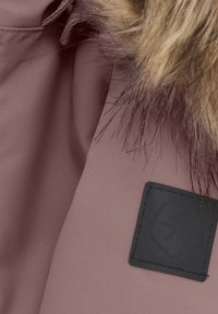 Rosa tyg med en slät textur, med en brun pälsdetalj och en svart rektangulär lapp med en upphöjd logotyp.