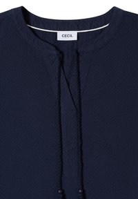 Chemise texturée bleu marine avec col rond, patte de boutonnage partielle sur le devant, liens tressés décoratifs terminés par des perles, et étiquette "CECIL" à l'intérieur du col.