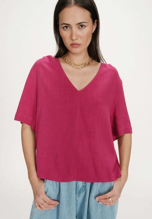 Femme portant un haut ample à col en V magenta, un jean bleu clair et des colliers à chaînes dorées superposées, debout les mains dans les poches.