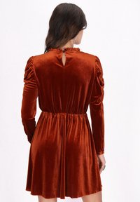 Rozsdabarna bársonydressz hosszú ujjakkal, redőzött vállakkal, szűkített derékkal és kulcslyuk hátkivágással. Sima textúra és harangszoknya.