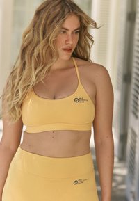 Sárga sportmelltartó és magas derekú leggings rugalmas anyagból, mindkét darabon logóval. Simarészű, minimális varrásokkal.