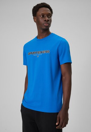 Mężczyzna w jaskrawo niebieskiej koszulce z napisem "SPORTSWEAR", stoi z rękami w kieszeniach, patrząc w bok na jednolitym tle.
