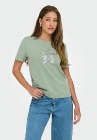 Lys grøn kortærmet T-shirt med et sølvfarvet pailletstjernedesign på forsiden, parret med højtaljede blå denim jeans.