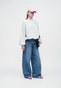 Mujer vestida con un sudadera gris claro, pantalones vaqueros de pierna ancha azules, zapatillas de deporte rosas y una gorra rosa, de pie contra un fondo blanco sencillo.