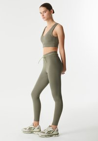 Soutien-gorge de sport vert olive et leggings taille haute assortis, avec un cordon de serrage et un matériau lisse et extensible. Portés avec des baskets grises.