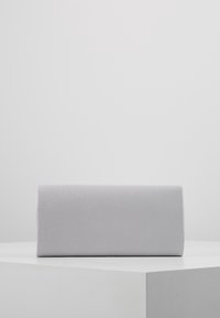 Pochette rectangulaire argentée à texture lisse posée sur une surface blanche devant un fond gris clair uni.