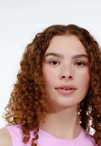 Ung person med lockigt, rödbrunt hår som bär en ljusrosa ärmlös topp. Klar hud med fräknar, neutral uttryck, mjuka drag.