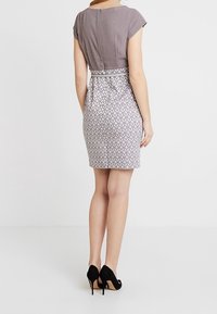 Grå kjole med tætsiddende overdel og mønstret skørt; har kappelignende ærmer, høj halsudskæring og lynlås bagpå. Båret med sorte hæle.