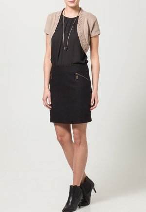 Cárdigan de punto beige sobre una blusa negra y una falda lápiz negra con dos bolsillos con cremallera. La modelo lleva botas negras hasta el tobillo.