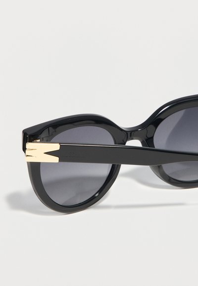Μαύρα γυαλιά ηλίου σε σχήμα γατίσιου ματιού με σκουρόχρωμους φακούς και χρυσή μεταλλική λεπτομέρεια στον βραχίονα, σε λευκό φόντο.