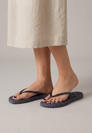Voeten met donkere gevlochten teenslippers en marineblauwe nagellak, staand op een lichte vloer, gedeeltelijk bedekt door een beige linnen rok.