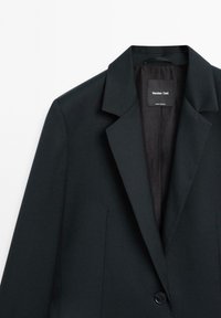 Černý blejzr s klopovým límcem s výřezem, jednoknalovým zapínáním a hladkou texturou. Uvnitř je štítek s názvem značky.