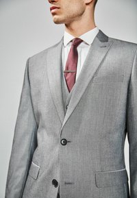 Harmaa muot tailored -takki, jossa on nokkalaput, kaksi mustaa nappia ja vasemman rinnan tasku. Mukana burgundinvärinen solmio ja valkoinen paita.