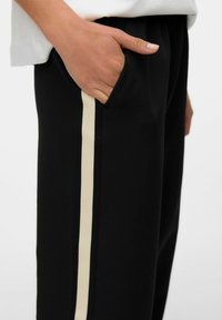 Zwarte broek met een strakke uitstraling en een ivoorkleurige streep aan de zijkant. Het materiaal lijkt glad, met zijzakken voor functionaliteit.