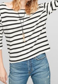 Gestreiftes Langarmoberteil in Schwarz und Weiß mit weitem Ausschnitt, kombiniert mit ausgebleichten blauen Jeans. Dazu gehört eine Halskette mit einem Anhänger und verschiedenen Charms.