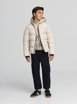 Giacca imbottita beige con cappuccio, dotata di cerniera nera e polsini a contrasto; indossata sopra una felpa crema con motivo a fiamma. Pantaloni cargo neri e scarpe da ginnastica.