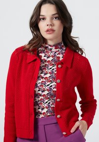Cardigan rosso lavorato a maglia con design a rete, colletto e bottoni argentati, indossato sopra una maglietta a fantasia floreale.