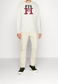 Sudadera blanca con logo rojo y marino, combinada con pantalones beige claros y zapatillas negras. La sudadera presenta un bolsillo canguro en el frente y cordones.