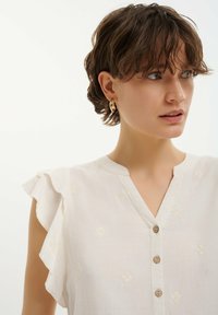 Top in lino bianco con maniche a sbuffo, chiusura con bottoni in legno e delicati motivi floreali ricamati. Presenta un design con scollatura a V.
