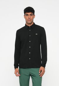 Maglietta nera a maniche lunghe con chiusura a bottoni, colletto a punta e logo ricamato in verde. Realizzata in tessuto morbido con una texture liscia.