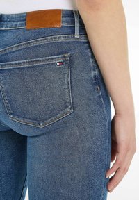 Jeans de denim azul con un acabado texturizado, que presentan una etiqueta de cuero marrón en la cintura y un pequeño logo rojo, blanco y azul en el bolsillo trasero.