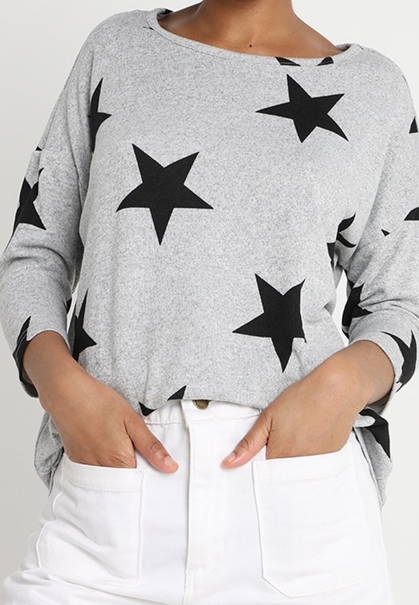 Personne portant un t-shirt gris à manches longues avec de grandes étoiles noires et un pantalon blanc, les mains dans les poches avant.