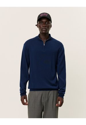 Navy blue zip-up sweater features a high collar and long sleeves, paired with gray plaid trousers. Black cap with pink text reads "Late Night Service."
