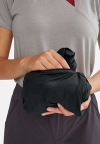 Bundles de tissu noir et froissé tenus dans une main, suggérant un design compact. T-shirt gris porté, avec une touche de rouge en dessous.