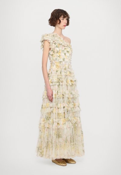 Een gelaagde, één-schouder jurk van doorschijnende stof, met ruffles en een bloemenspatroon in pastelkleuren, gecombineerd met gouden platte schoenen.