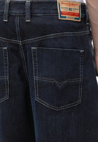 Σκούρο μπλε τζιν από denim, πίσω όψη, που δείχνει δύο τσέπες με μπεζ ραφές και ορθογώνια δερμάτινη ετικέτα με το λογότυπο στη μέση.