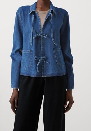 Persona con una chaqueta de mezclilla azul con bolsillos frontales y cierres de lazo, sobre una camiseta beige y pantalones negros, de pie frente a un fondo liso.