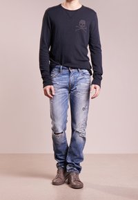 Tmavě modrá košile s dlouhým rukávem s motivem lebky a zkřížených kostí, doplněná o seprané světle modré džíny a šedé tenisky s vysokým střihem.