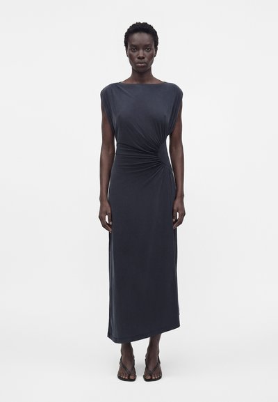 Femme portant une robe maxi sans manches gris foncé avec fronces sur les côtés, debout droite devant un fond clair uni, portant des sandales noires.