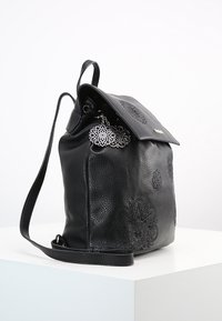 Schwarzer Leder Rucksack mit geprägten Blumenmustern, umschlagbarem Deckel, Metallanhänger und verstellbarem Träger. Strukturiertes Finish.