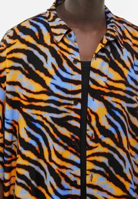 Koszula z krótkim rękawem wykonana z przezroczystej tkaniny, zdobiona czarnym, pomarańczowym i niebieskim wzorem pasków tygrysa, z zapinanym na guziki przodem i klasycznym kołnierzykiem.