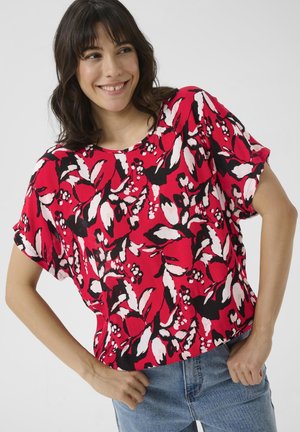 Blouse florale rouge à coupe large, avec des manches courtes retroussées et des accents noirs. Le tissu présente un motif texturé avec des feuilles blanches et des baies.