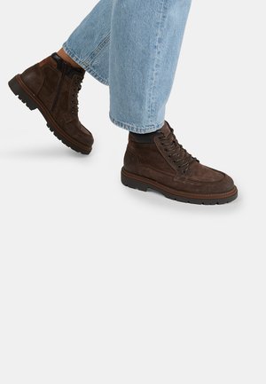 Persona vestida con botas de cuero gamuza marrones con cordones y cremallera lateral, y jeans de mezclilla azul claro, sobre fondo blanco.
