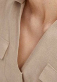 Blazer beige con solapas de muesca y bolsillos tipo parche. Textura de tela suave, con un escote profundo que revela parte del hombro.