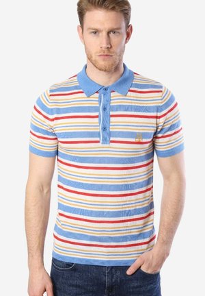 Mann trägt ein kurzärmeliges Poloshirt mit blauen, roten und gelben horizontalen Streifen und dunkle Jeans, steht mit einer Hand in der Tasche.