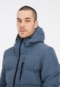 Blauer Pufferjacke mit Kapuze, ausgestattet mit einem Reißverschluss vorne, verstellbaren Kordelzügen und genähten Paneelen zur Isolierung.