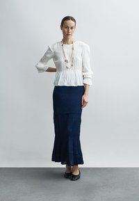Witte geplooide blouse met driekwart mouwen, gecombineerd met een marineblauwe, gestructureerde maxi-rok met een uitlopende zoom en eenvoudige zwarte schoenen.
